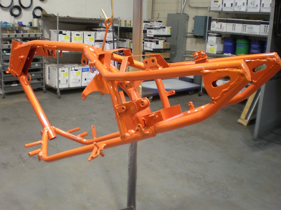An orange motorcycle frame is being powder coated in a workshop.